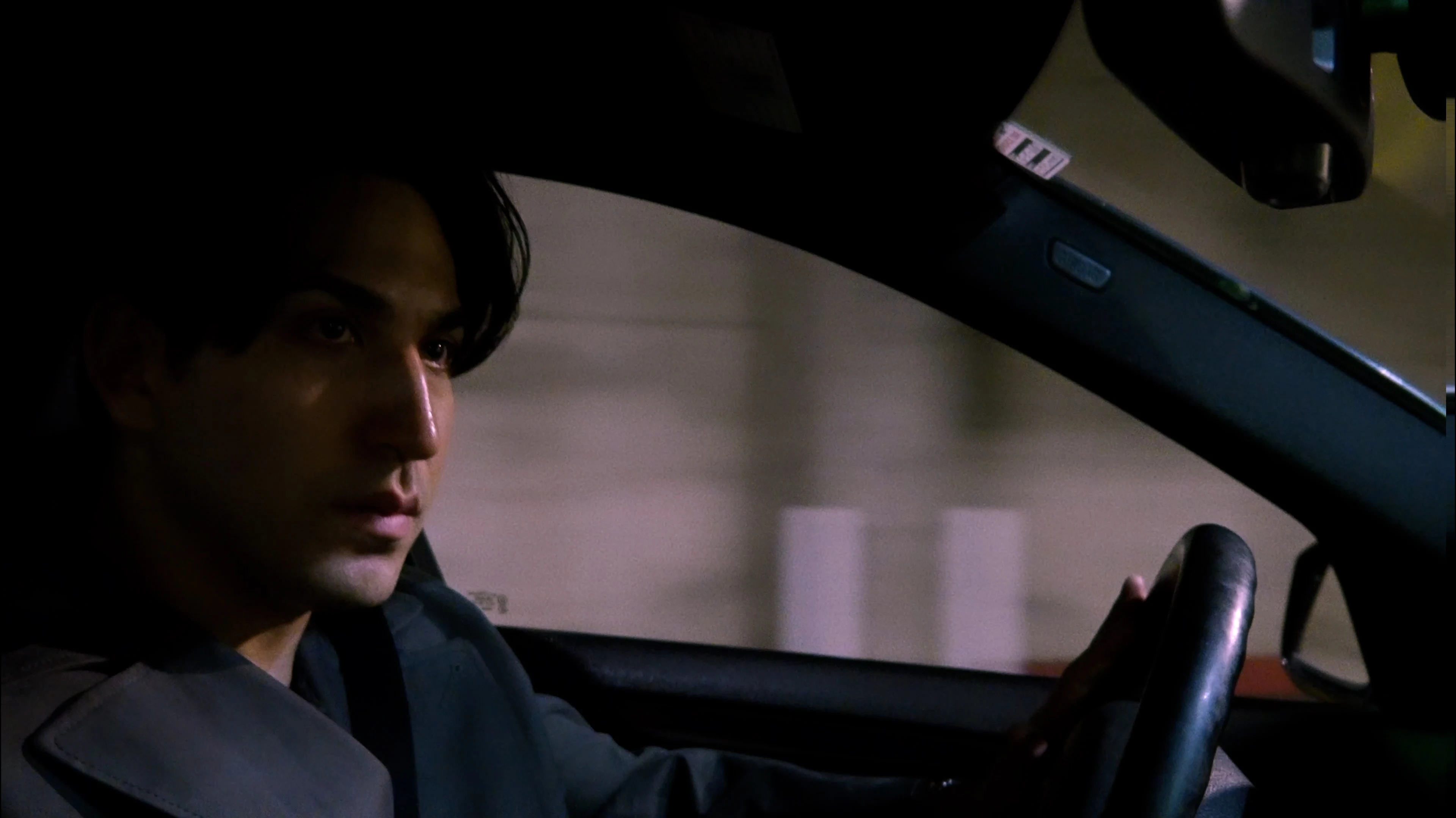 Ian Bloom in King standing in profile in a concrete garage wearing a trench coat